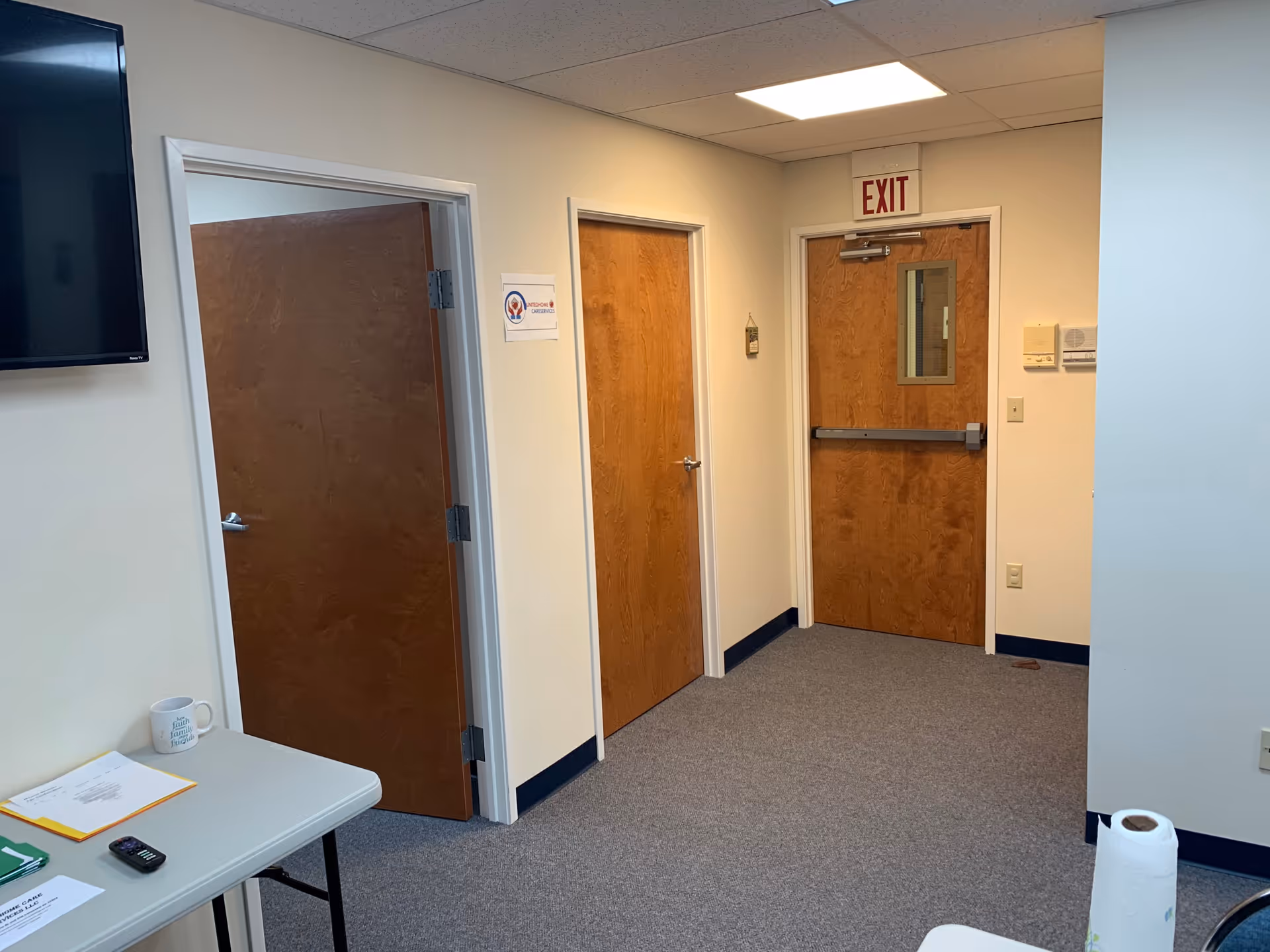 Interior hallway in a senior facility with wooden doors, exit sign, and a table with papers.