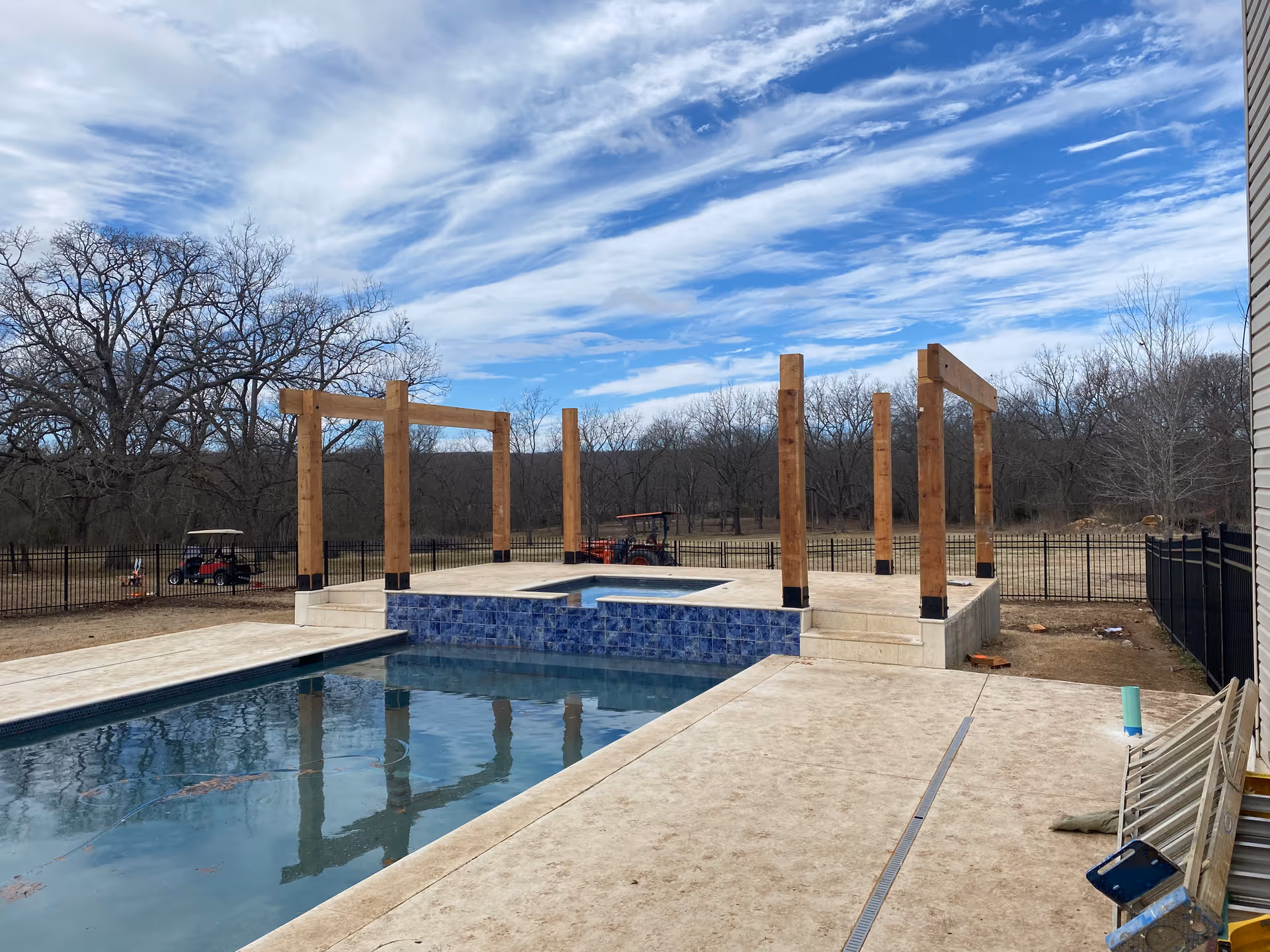 Outdoor pool area under construction with tall wooden posts and blue-tile raised spa.