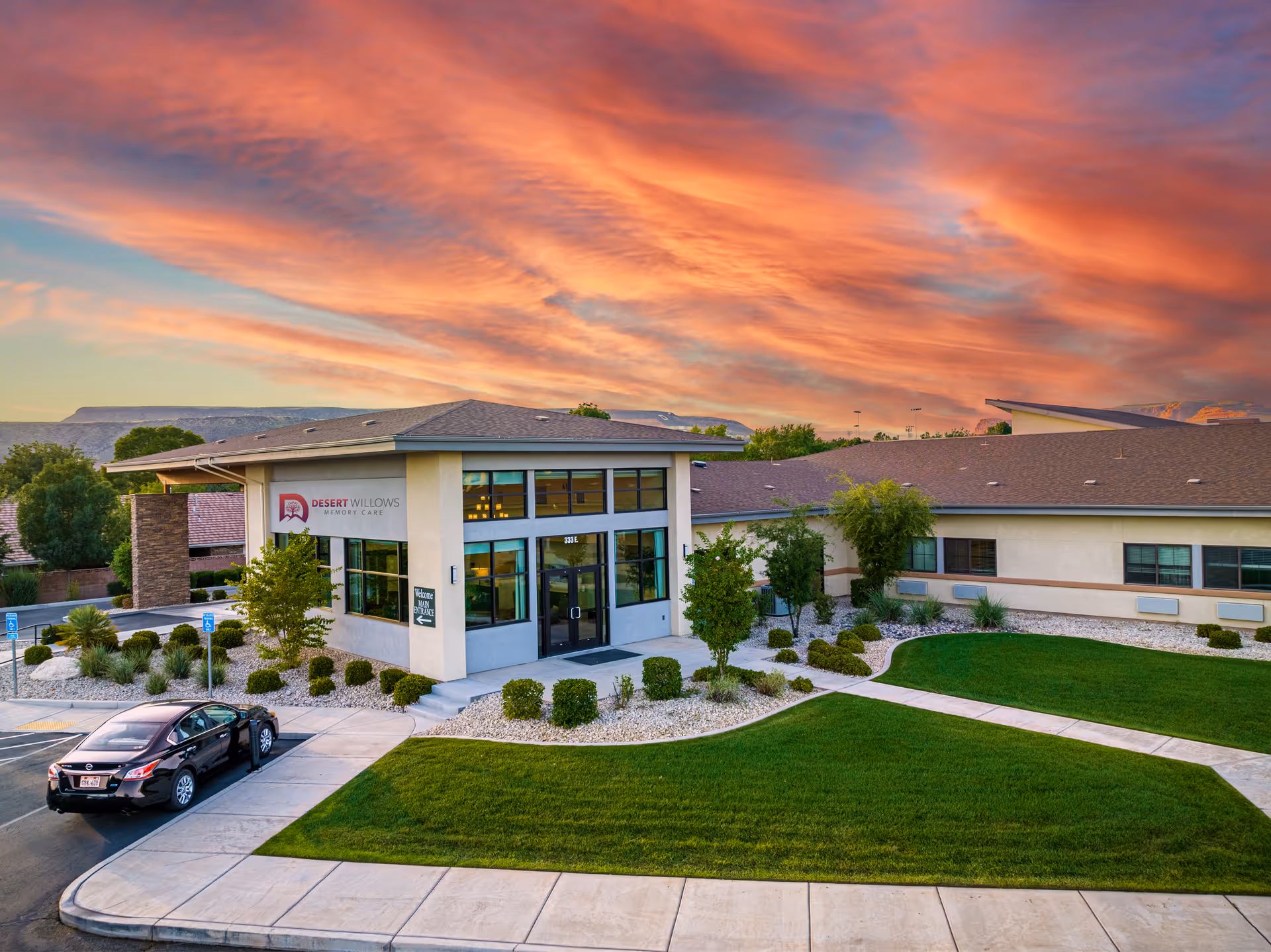 A modern senior living facility exterior with glass entry, manicured lawn, and parking.