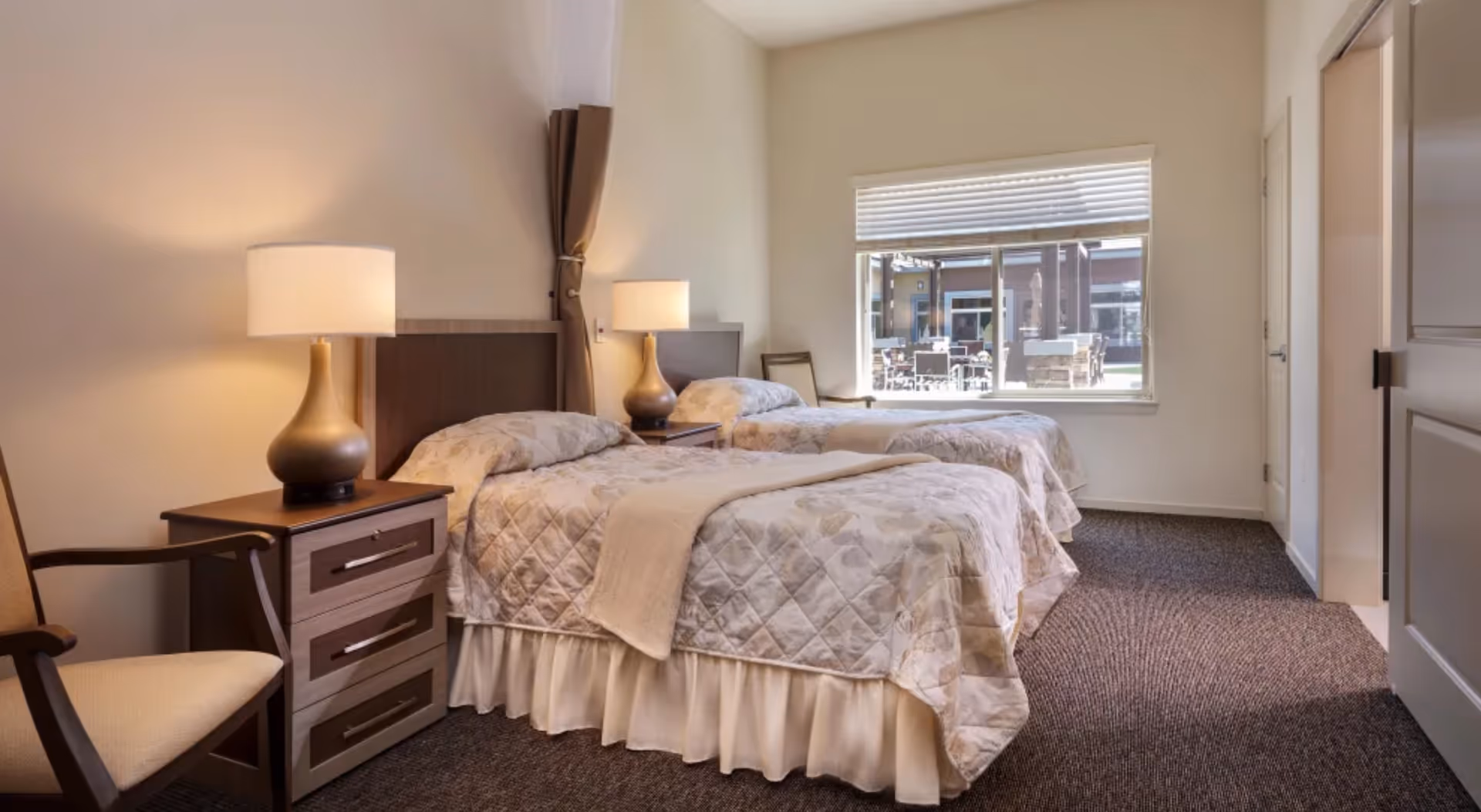 Bright bedroom with two twin beds, bedside tables and lamps, a chair, and a window overlooking an outdoor patio.