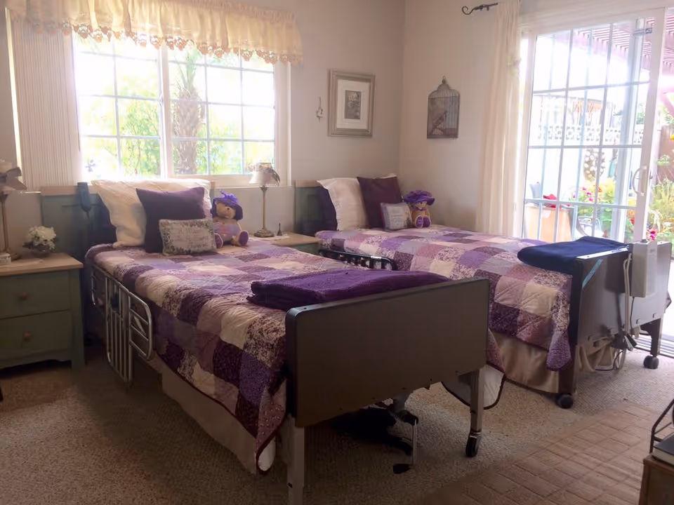 A cozy bedroom in The Hills of Hayward facility featuring two hospital-style beds with purple and white patchwork quilts, each adorned with pillows and a stuffed bear wearing a purple hat. The room has a large window with sheer curtains letting in natural light and a sliding glass door leading to an outdoor area. There are nightstands with lamps and decorative items beside each bed.
