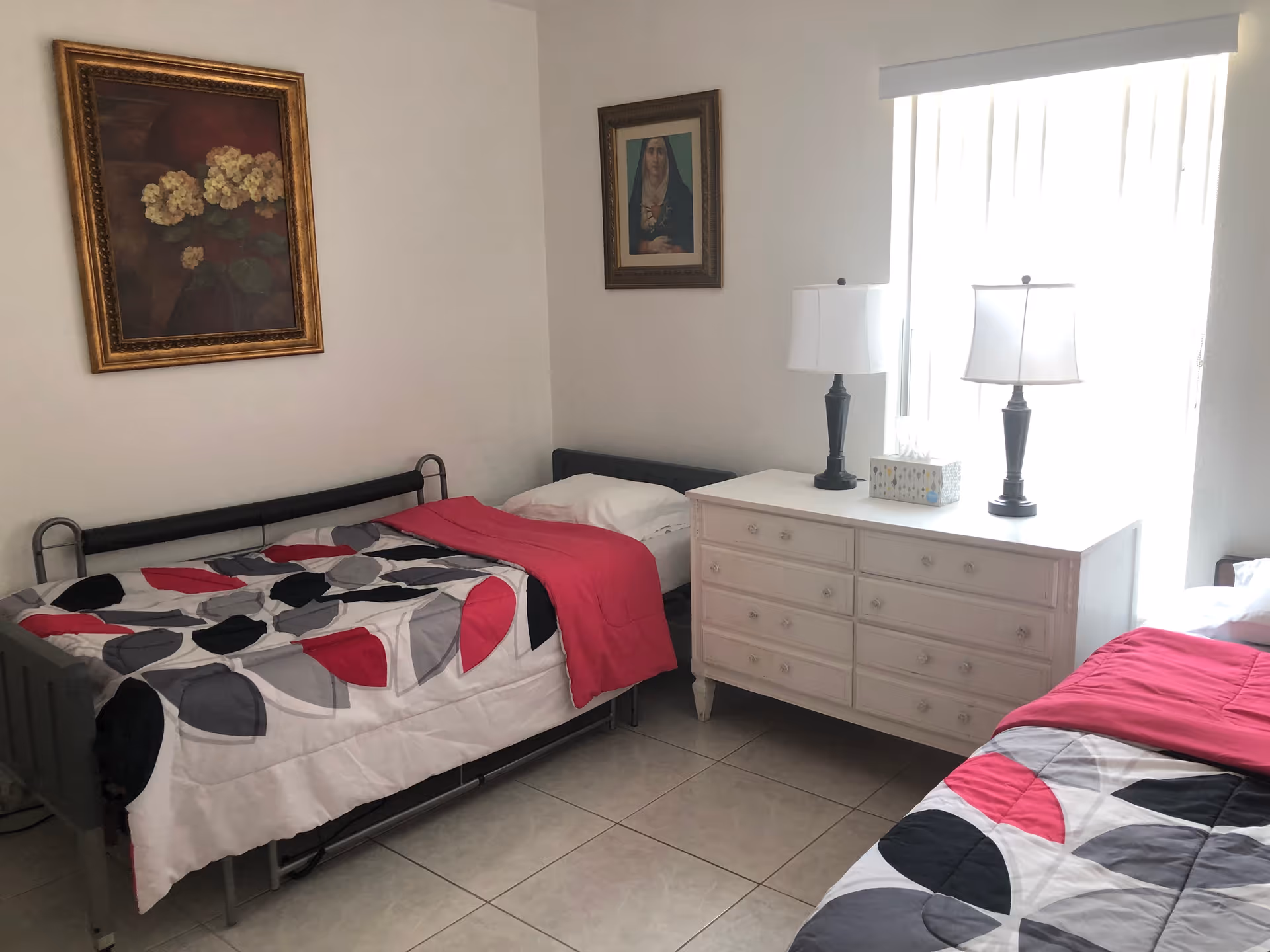 A small bedroom with two single beds, each with a colorful bedspread featuring red, black, gray, and white patterns. Between the beds is a white dresser with six drawers, topped with two black table lamps and a box of tissues. The room has tiled floors and white walls adorned with two framed paintings, one of flowers and the other of a woman. A window with vertical blinds allows natural light into the room.
