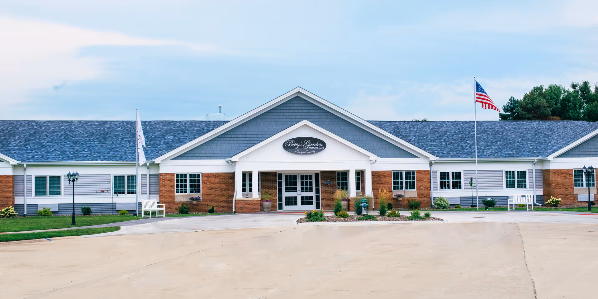 Front exterior view of a single-story senior living facility building with a peaked roof, brick and siding facade, two flagpoles with flags, benches, and landscaped greenery in front.