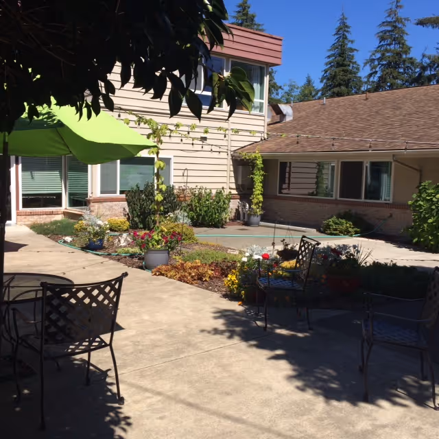 Outdoor courtyard area at a senior living facility with patio chairs, a green umbrella, flower beds, and a beige building with windows surrounding the courtyard. Tall evergreen trees are visible in the background under a clear blue sky.