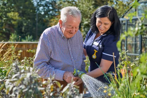 An elderly man and caregiver garden outdoors, watering plants in a sunny facility garden.