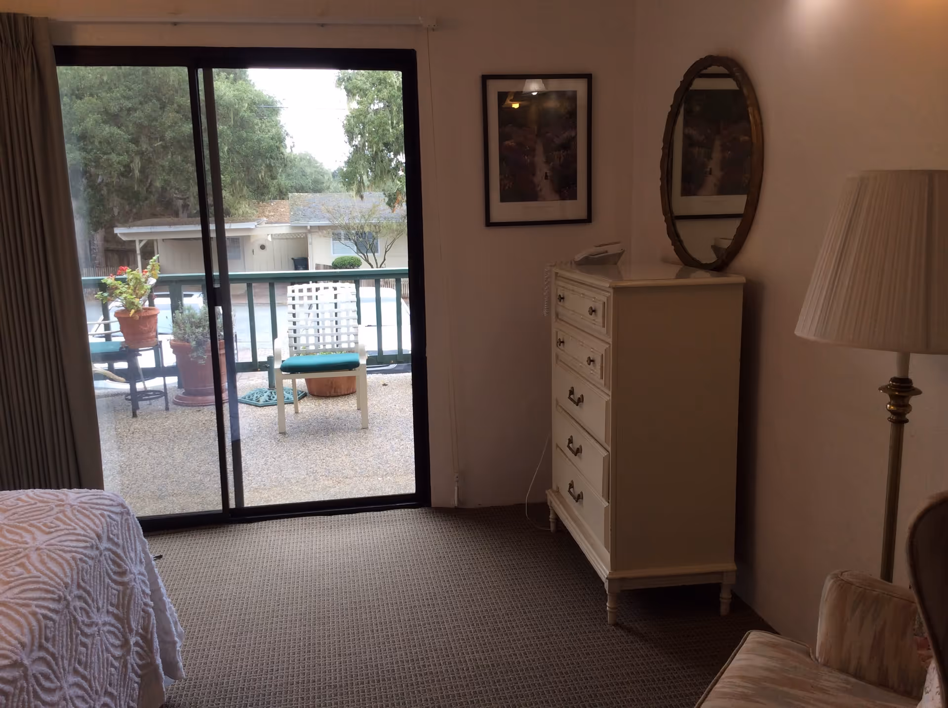 A bedroom with a sliding glass door leading to an outdoor patio. Inside the room, there is a white dresser with a telephone on top, a round mirror hanging above it, a framed picture on the wall, a floor lamp, and a cushioned chair. Outside on the patio, there are potted plants and a white chair with a green cushion. Trees and a neighboring building are visible in the background.