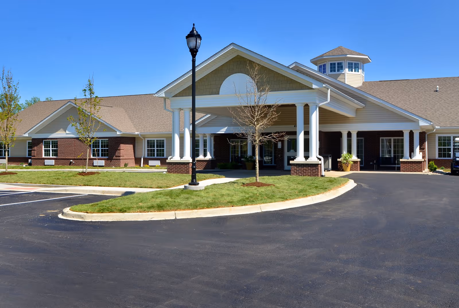 Exterior view of The Springs of Lima senior living facility showing a large building with a covered entrance supported by white columns, a brick and beige siding facade, a small tree and lamp post in front, and a clear blue sky.
