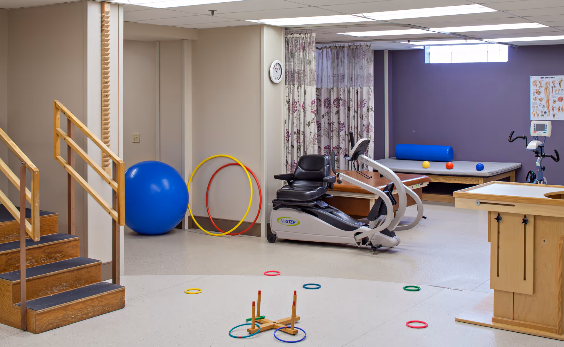 A physical therapy or rehabilitation room with exercise equipment including a recumbent exercise bike, large blue exercise ball, colorful hoops, a wooden ring toss game, and a padded therapy table against a purple wall with anatomical charts.