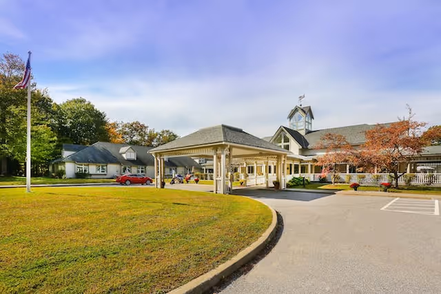 Front entrance of a senior living facility with a covered porte-cochere, driveway, lawn, and flagpole.