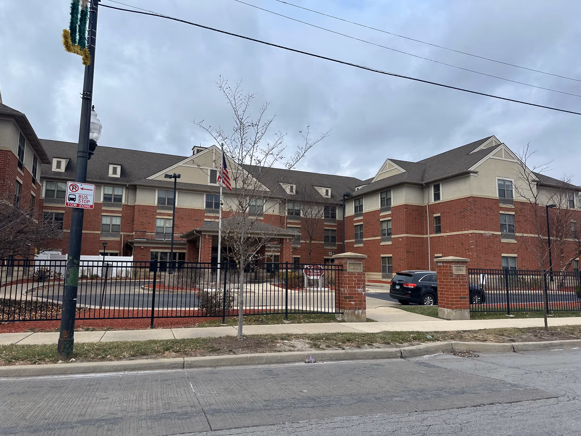 Exterior view of a three-story brick and beige building with multiple windows, a black metal fence, and a driveway entrance. There is a flagpole with an American flag near the entrance and a black SUV parked inside the fenced area. The sky is cloudy and there is a street with a sidewalk in the foreground.