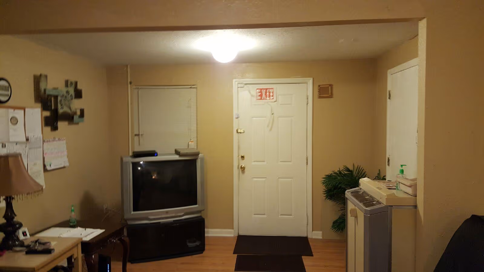 Cozy living area with TV on a stand, exit door, desk, lamp, and a laundry appliance.
