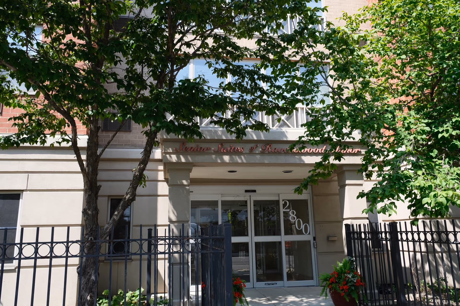 Entrance of the Senior Suites of Ravenswood Manor building with glass doors marked 2800, flanked by trees and a metal fence.