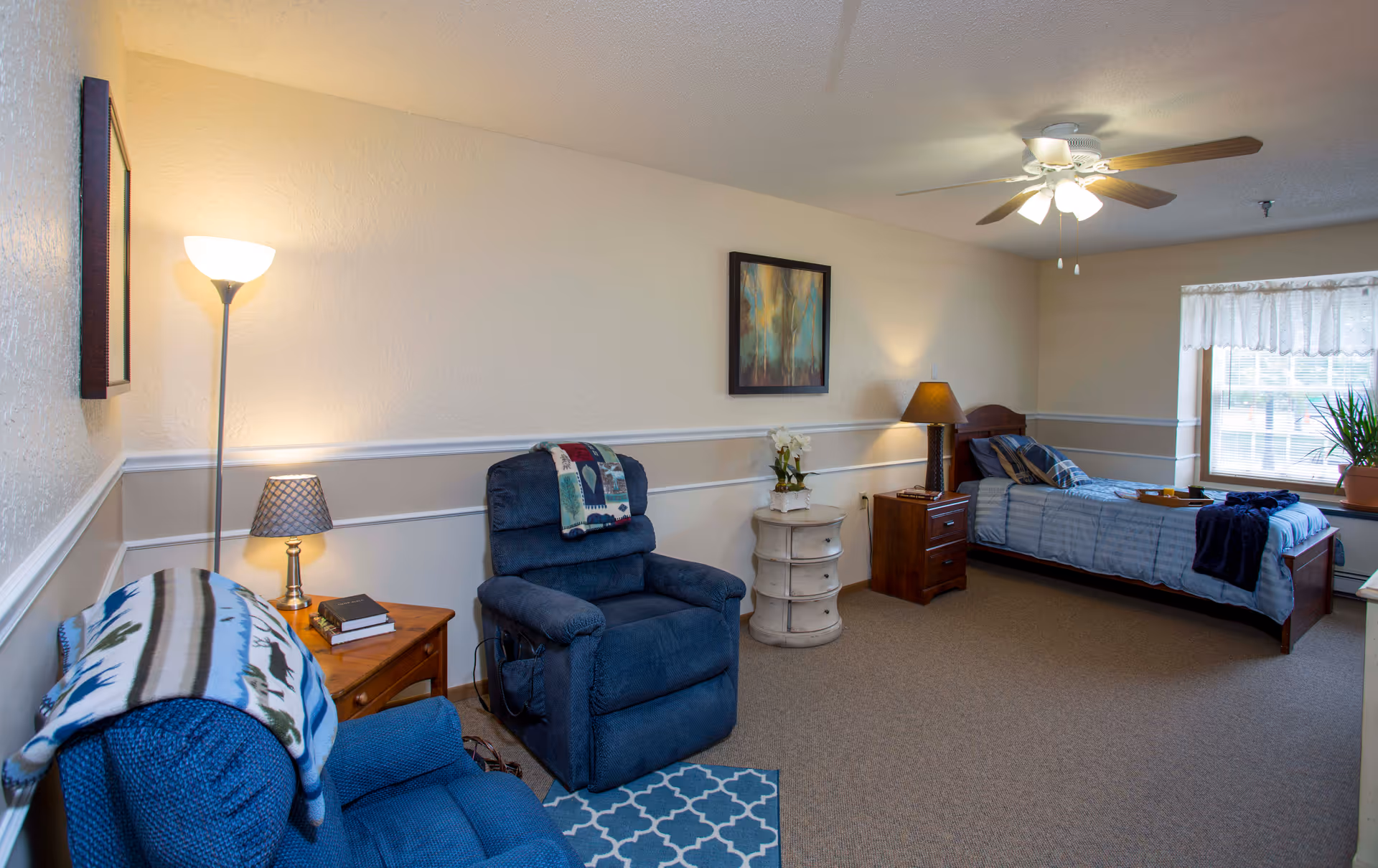 A furnished bedroom in a senior facility with a bed, recliners, and lamps.