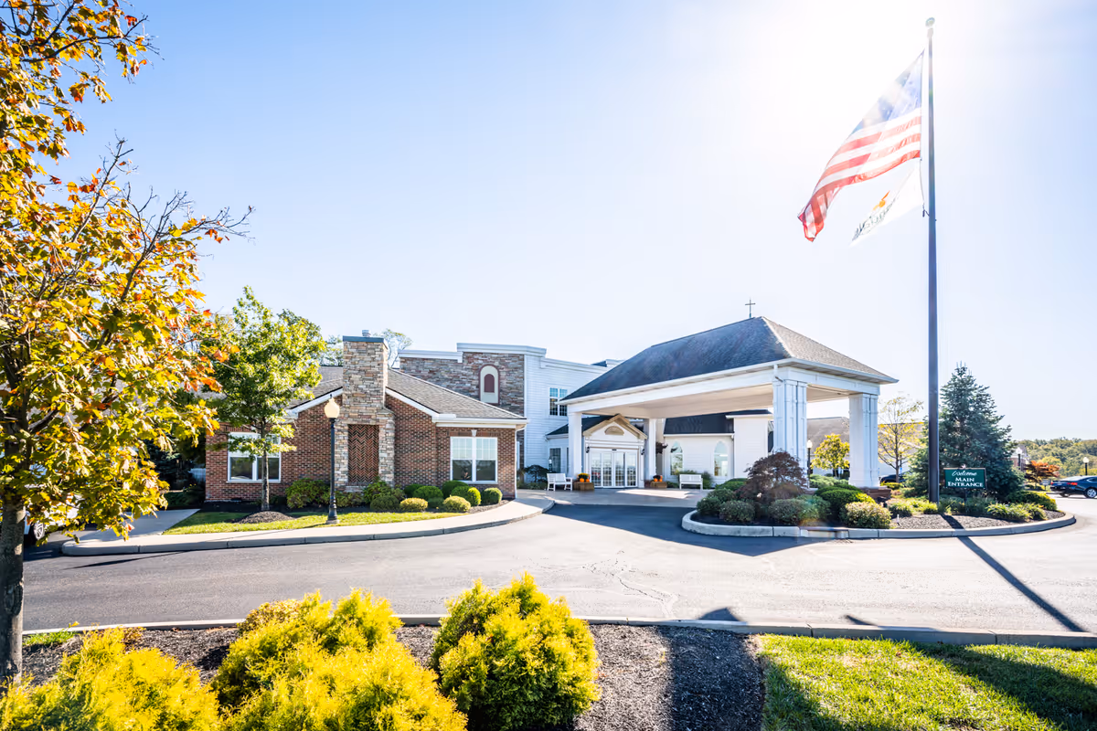 Front entrance of a brick senior living facility with a covered porte-cochere, landscaped driveway, and an American flag flying on a tall pole.