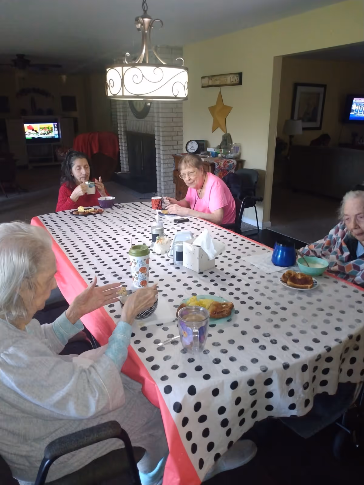 Four elderly women sitting around a rectangular dining table covered with a white tablecloth with black polka dots and a pink border, eating and drinking. The room has a hanging light fixture above the table, a white brick fireplace in the background, and a wall decorated with a large yellow star and a sign that says 'LOVE'.