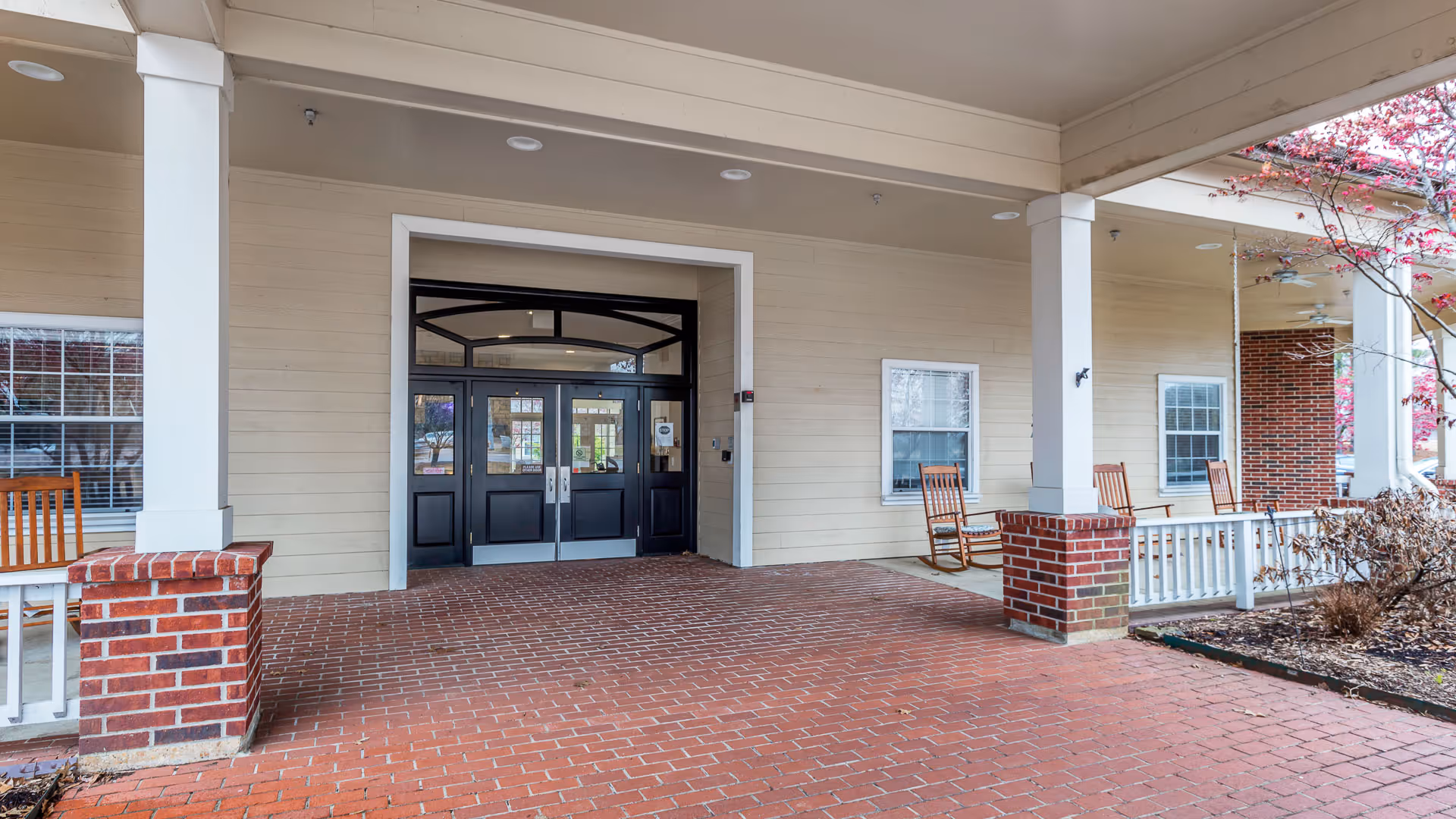 Covered entrance area of Hearthstone Assisted Living with double glass doors, brick pillars, and several wooden rocking chairs on the porch. There are windows on the beige siding walls and a tree with pink blossoms on the right side.