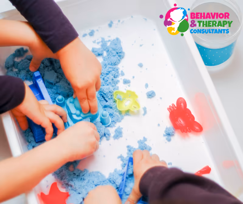 Two hands play with blue sensory sand and small toys in a tray.