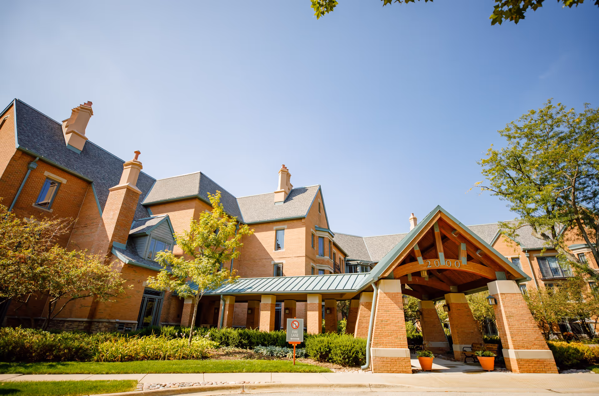 Exterior view of a large brick building with multiple chimneys and a covered entrance with the number 20000 displayed. The building is surrounded by green trees and shrubs under a clear blue sky.