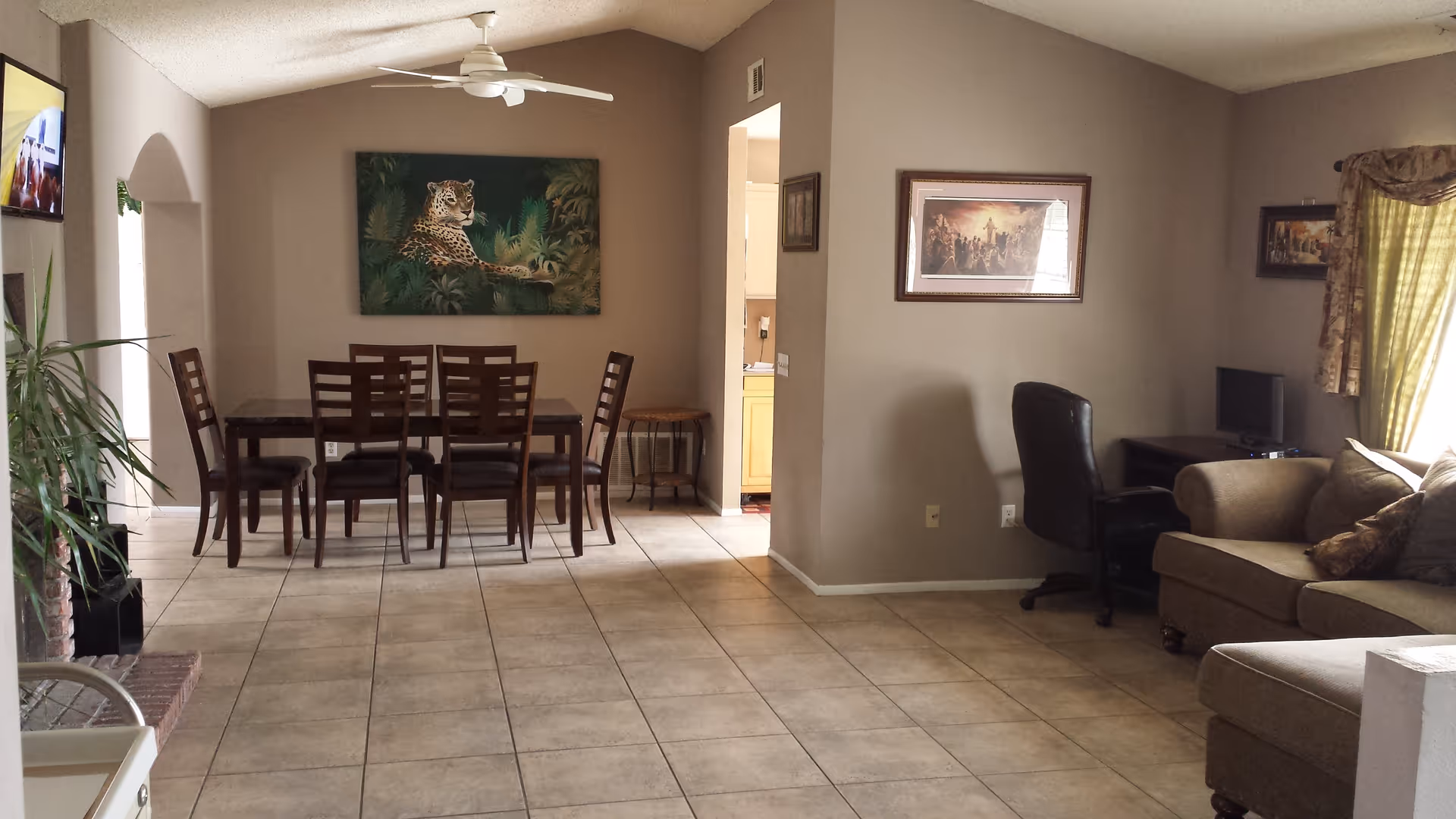 A spacious living and dining area with beige tiled floors and light brown walls. The dining area features a dark wooden table with six matching chairs and a large painting of a leopard on the wall. The living area has a beige sectional sofa with cushions, a black office chair, a small desk with a computer monitor, and framed artwork on the walls. A ceiling fan is mounted on the vaulted ceiling, and a window with patterned curtains allows natural light into the room.