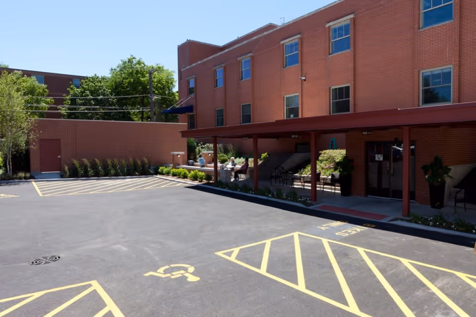 Exterior view of Aperion Care Wesley showing a brick building with multiple windows, a covered entrance area with chairs and plants, a parking lot with marked handicap parking spaces, and a few people sitting at a table near the entrance under a shaded area.
