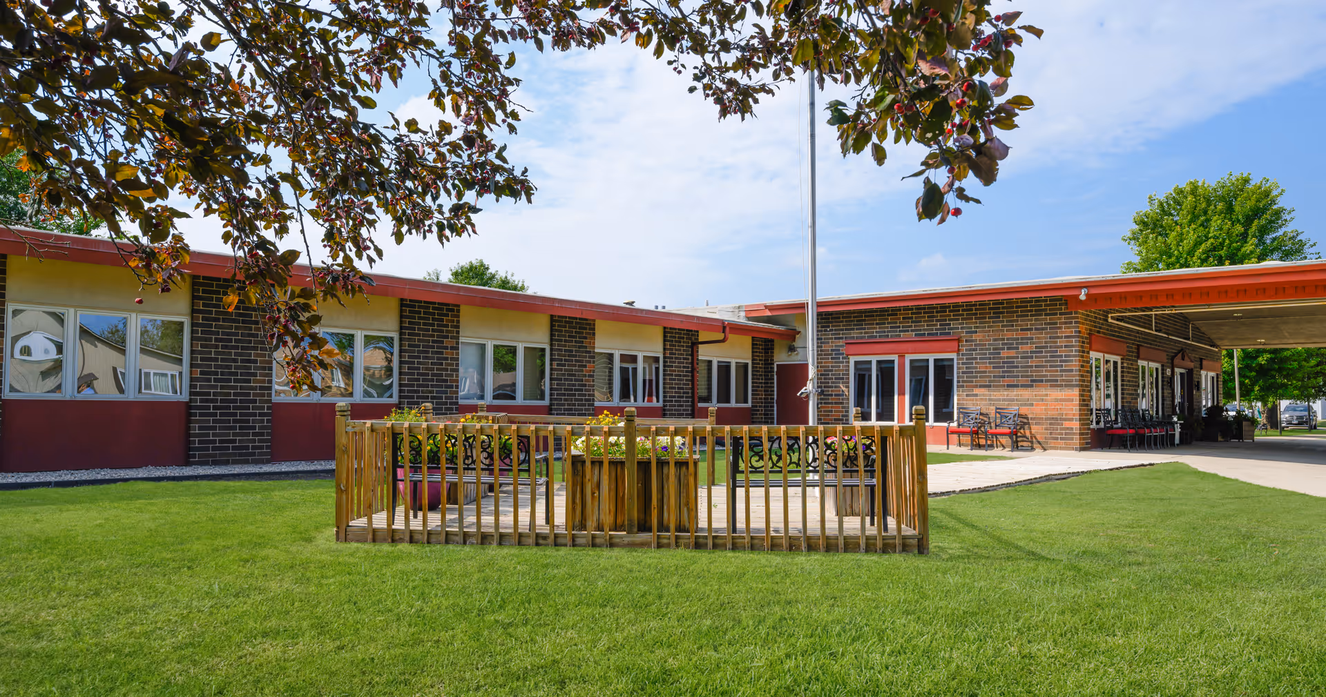 Single-story brick senior living facility with red trim, green lawn, and outdoor seating.