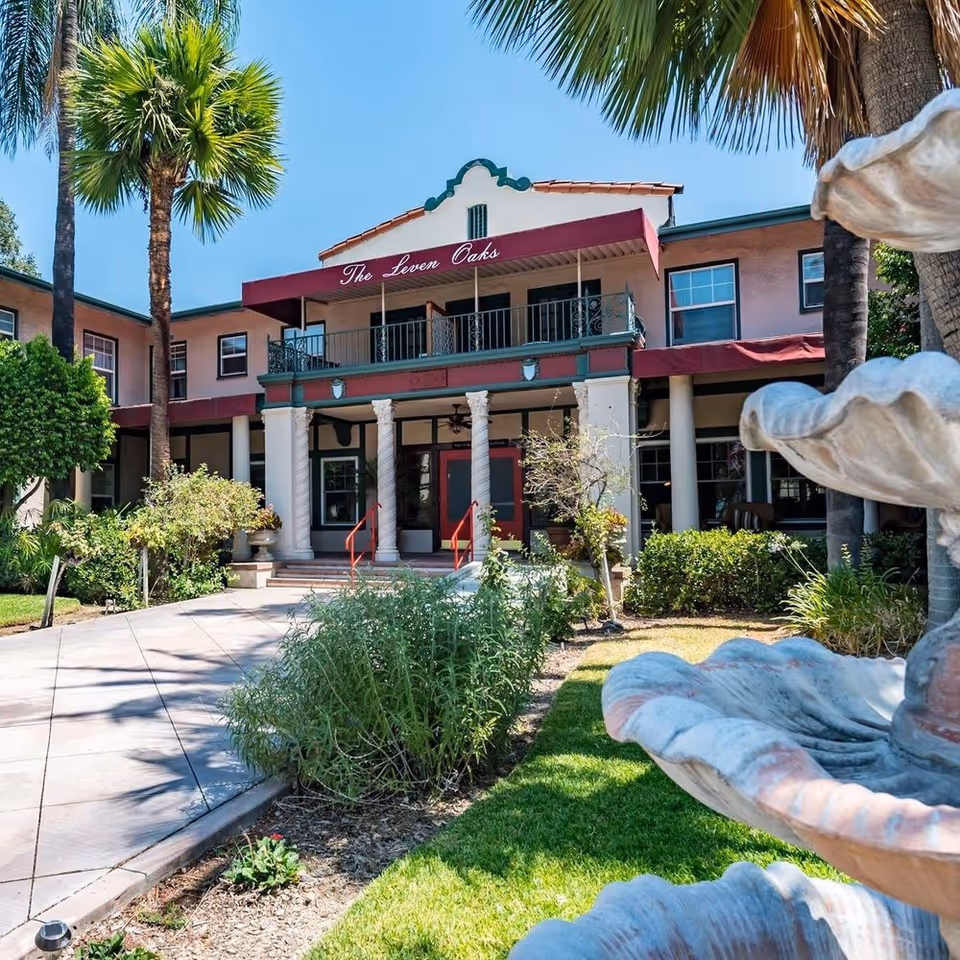 Exterior view of The Leven Oaks building at Henrietta's Leven Oaks by SCH, showing a two-story structure with a red awning, white columns at the entrance, palm trees, shrubs, and a multi-tiered stone fountain in the foreground under a clear blue sky.
