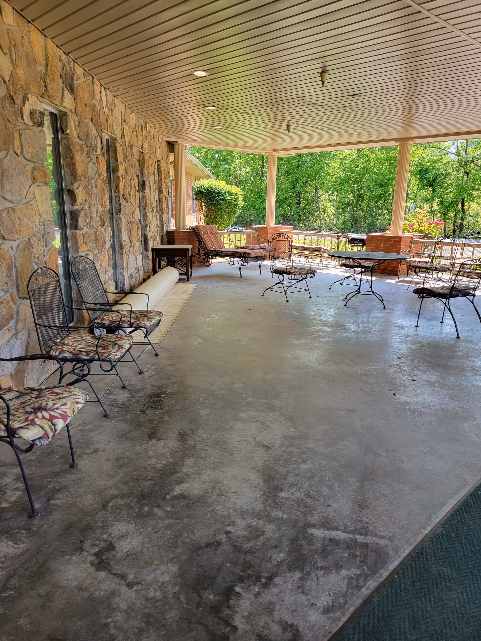 Covered outdoor patio area with stone wall on one side and several metal chairs with floral cushions, metal tables, and lounge chairs. The patio overlooks a green, wooded area with trees and bushes.