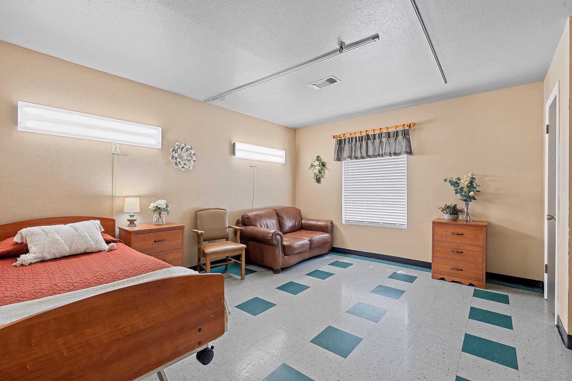 A nursing center bedroom with a wooden hospital bed covered in a red quilt and a white pillow. Next to the bed is a wooden nightstand with a small lamp and a vase of flowers. There is a brown leather loveseat and a wooden chair with armrests against the beige walls. The floor has a green and white checkered tile pattern. A window with closed blinds and a short curtain is on the far wall, with two wall-mounted lights above the bed area. A small wooden dresser with plants on top is near the window.