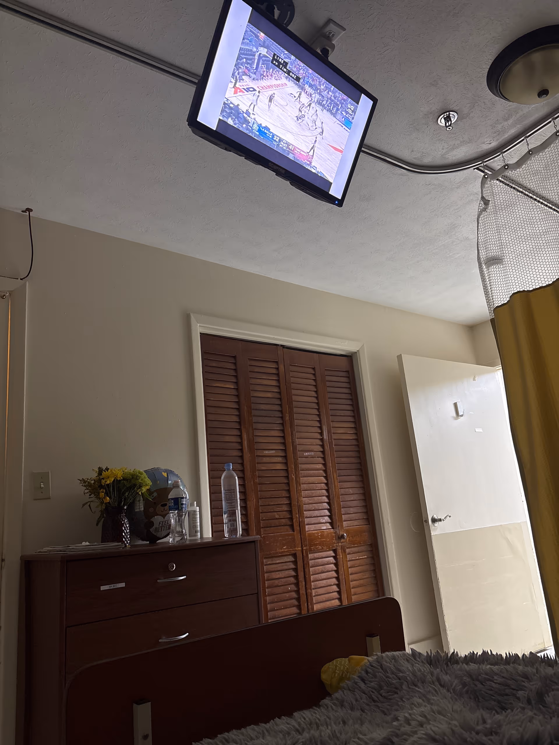 Senior living bedroom with ceiling-mounted TV, dresser, flowers, water bottle, and closed wooden closet.