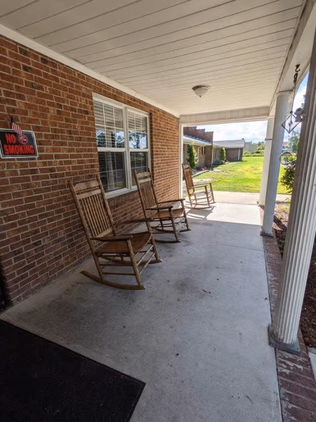 Covered brick front porch with three wooden rocking chairs and a 'No Smoking' sign overlooking a lawn.