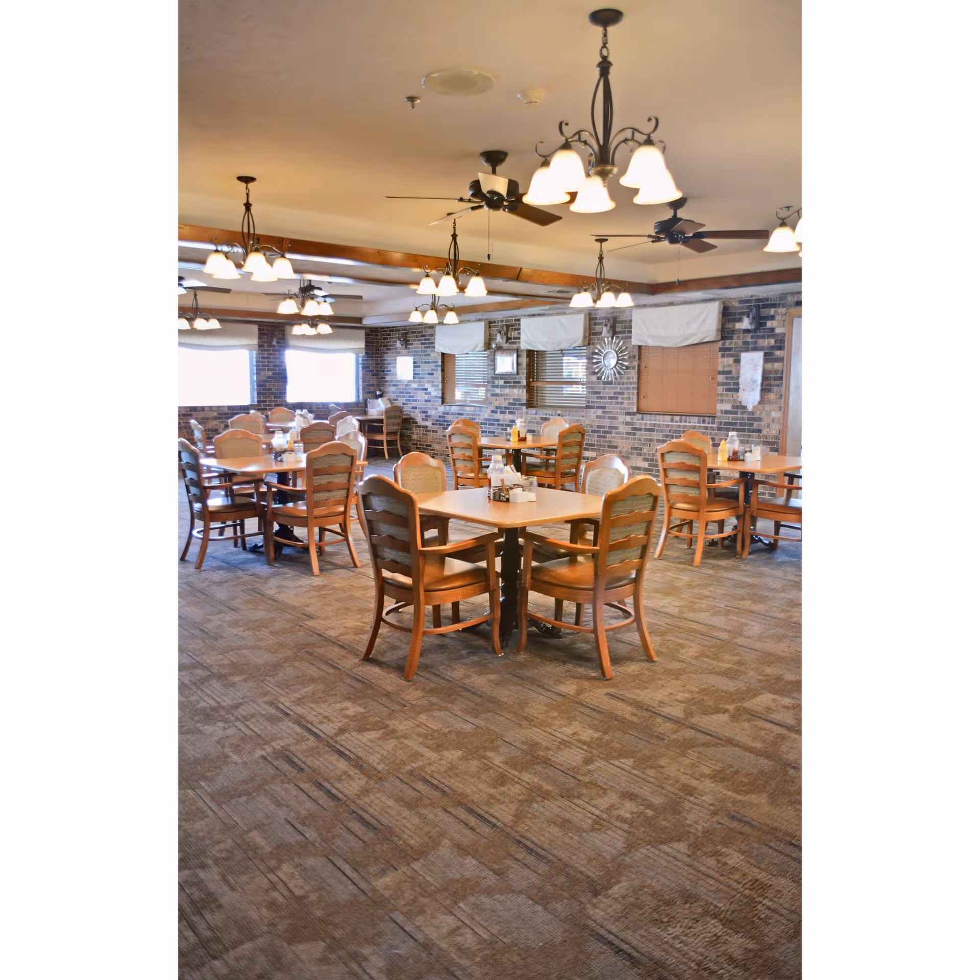A spacious dining room with multiple tables and wooden chairs, brick walls, chandeliers, and ceiling fans.