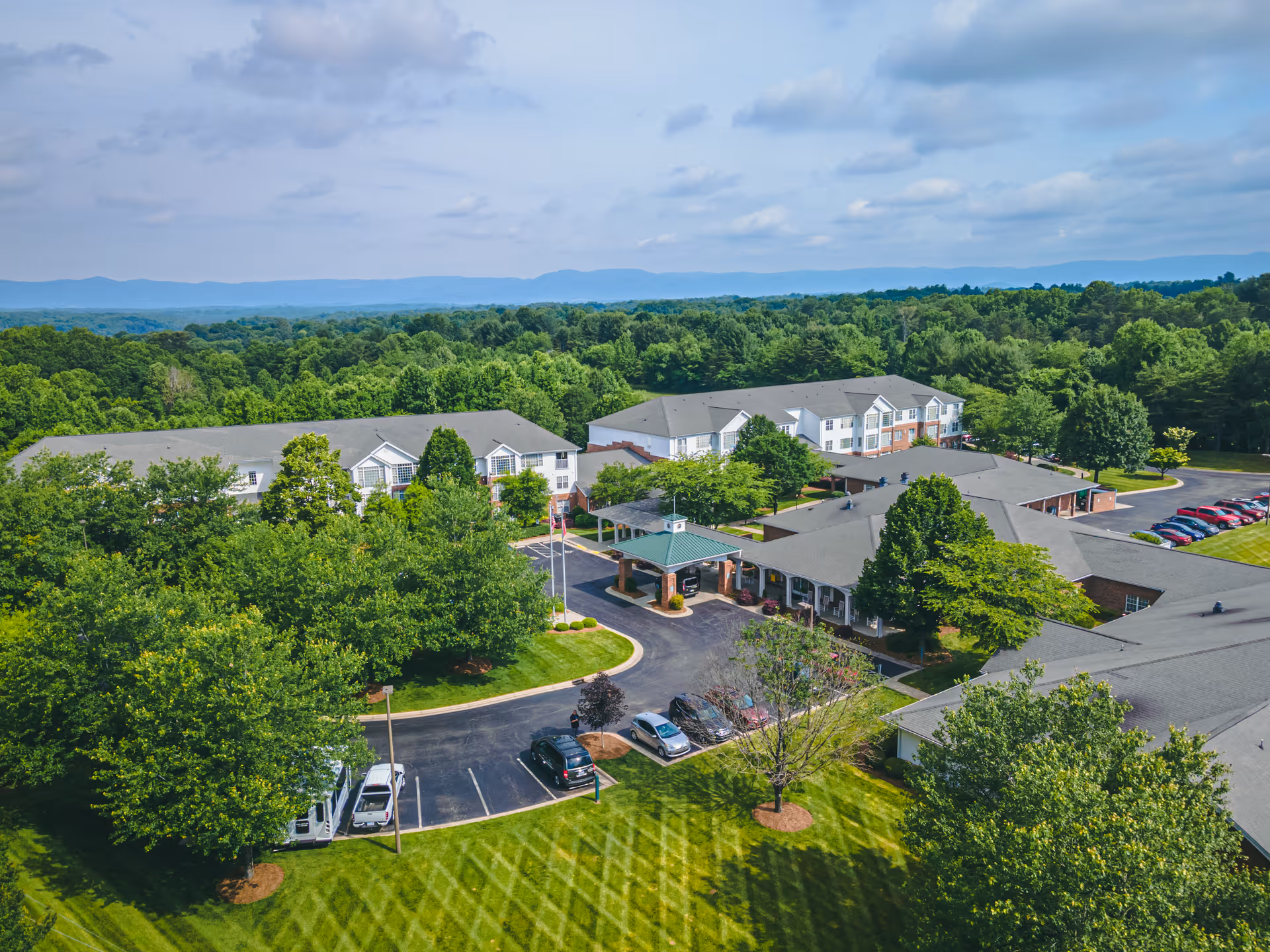 Aerial view of RidgeCrest senior living facility surrounded by lush green trees and landscaped lawns, with multiple connected buildings, a parking lot with cars, and a covered entrance area under a partly cloudy sky.