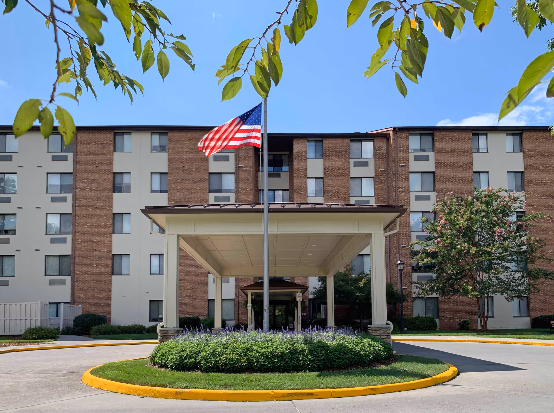 Front exterior view of a multi-story senior living facility named Edinburgh Square with a covered entrance, an American flag on a flagpole, and landscaped greenery including bushes and a flowering tree under a clear blue sky.
