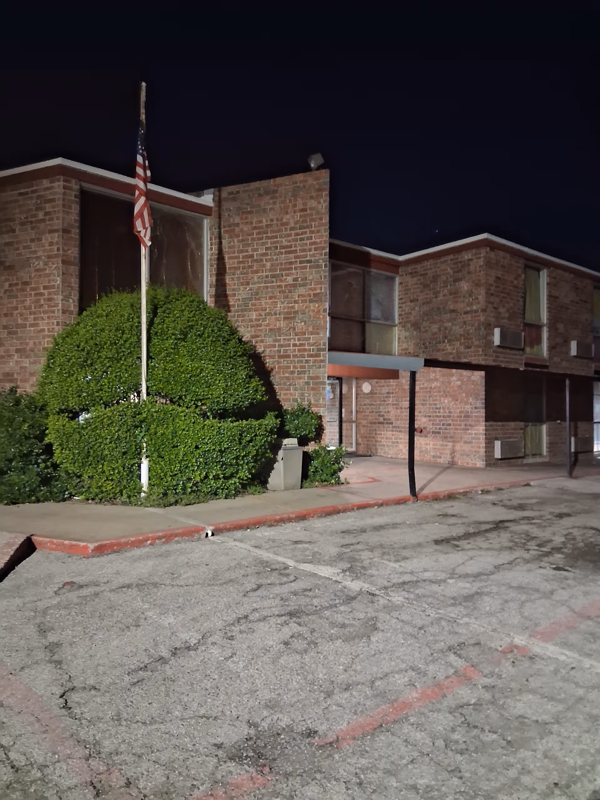 Night exterior of a brick senior living facility with flagpole and trimmed shrubbery.
