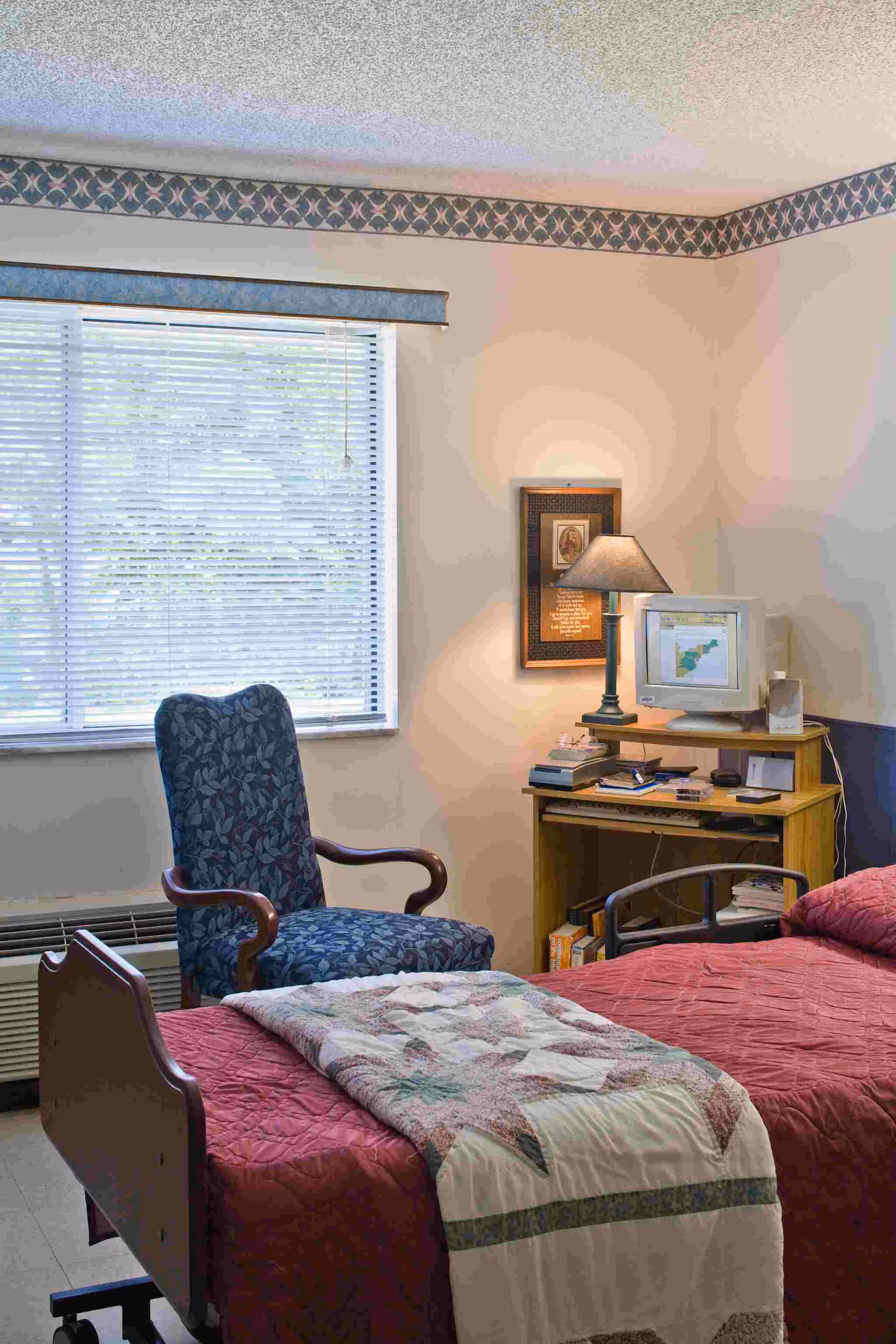 A cozy bedroom in a senior living facility featuring a single bed with a red quilt and a patterned blanket folded at the foot. Next to the bed is a blue upholstered armchair with wooden armrests. A wooden desk holds a vintage computer monitor, a lamp, and some books and papers. A window with blinds lets in natural light, and a framed picture hangs on the wall above the desk.
