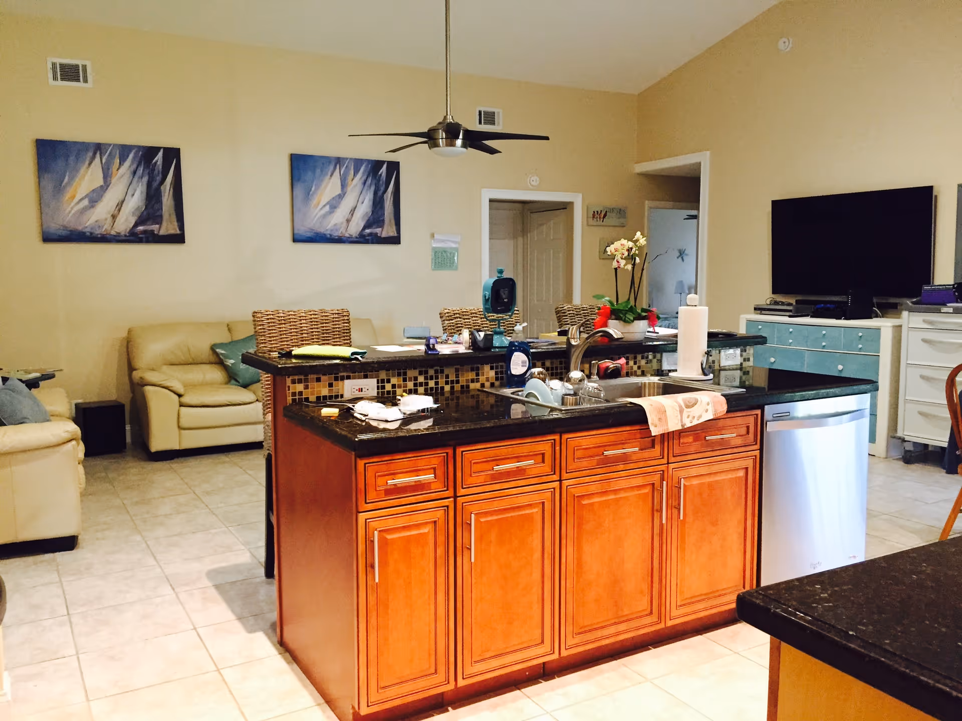 Interior view of a living space in an assisted living facility featuring a kitchen island with a sink, dishwasher, and various items on the counter. In the background, there are beige leather sofas, a ceiling fan, two paintings of sailboats on the wall, a flat-screen TV on a blue cabinet, and a dining area with wicker chairs.
