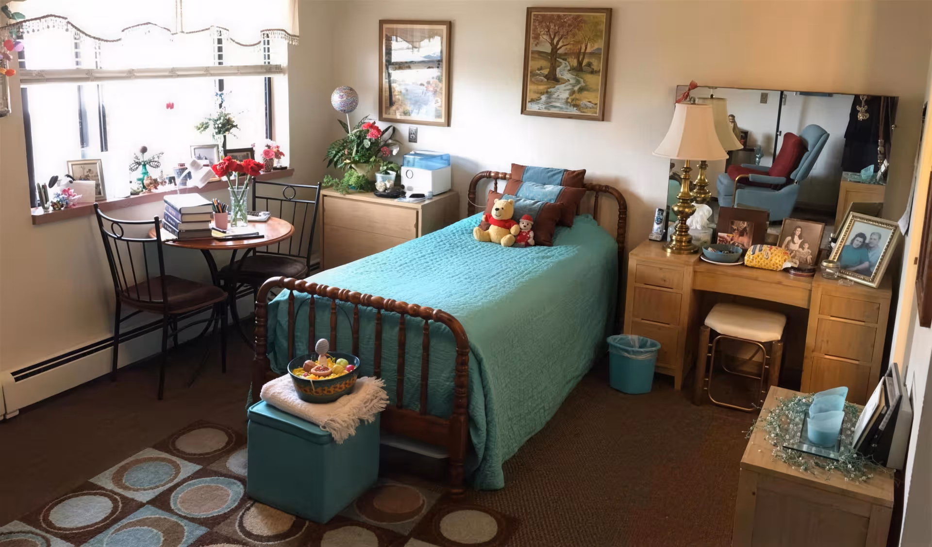 Cozy assisted-living bedroom with teal quilted bed, dresser, and window seating.