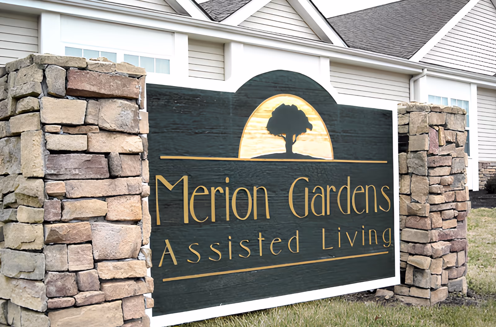 A large wooden sign reading "Merion Gardens Assisted Living" flanked by stone pillars in front of a light-colored building.
