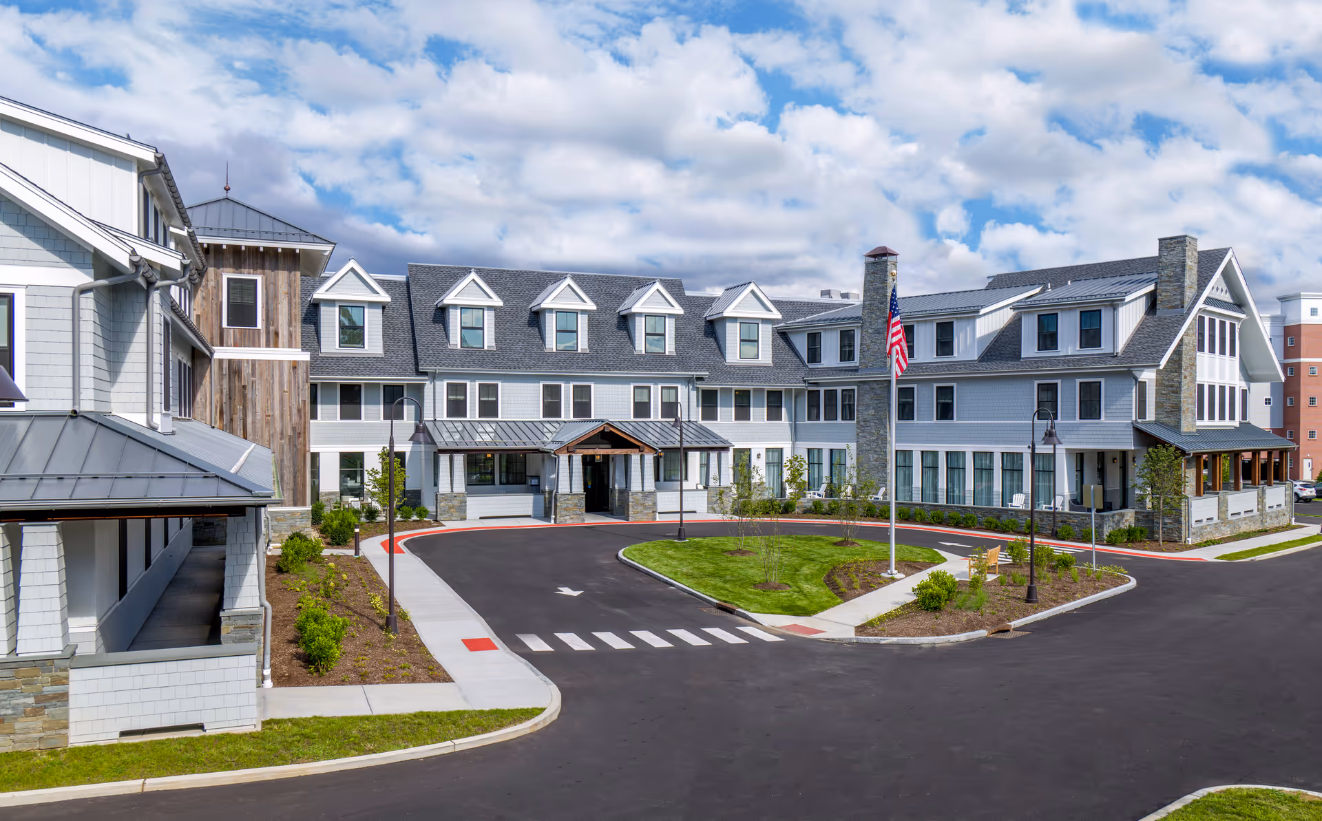 Exterior front view of a multi-story senior living residence with a covered main entrance, circular driveway, flagpole, and landscaped grounds.