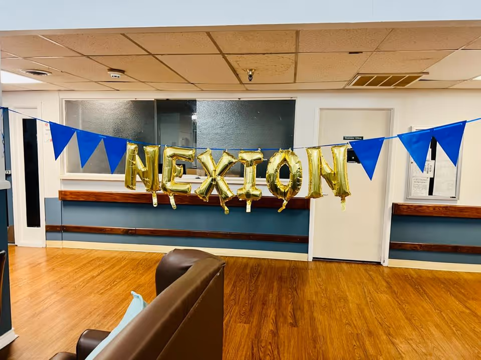 Interior view of a room with wooden flooring and a brown couch in the foreground. Blue triangular pennant banners and gold foil balloons spelling 'NEXION' are strung across the room. The walls are white with blue paneling and there is a door labeled 'Business Office' in the background.
