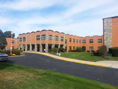 Brick senior living facility with curved windows, entrance, manicured lawn, and parking lot.
