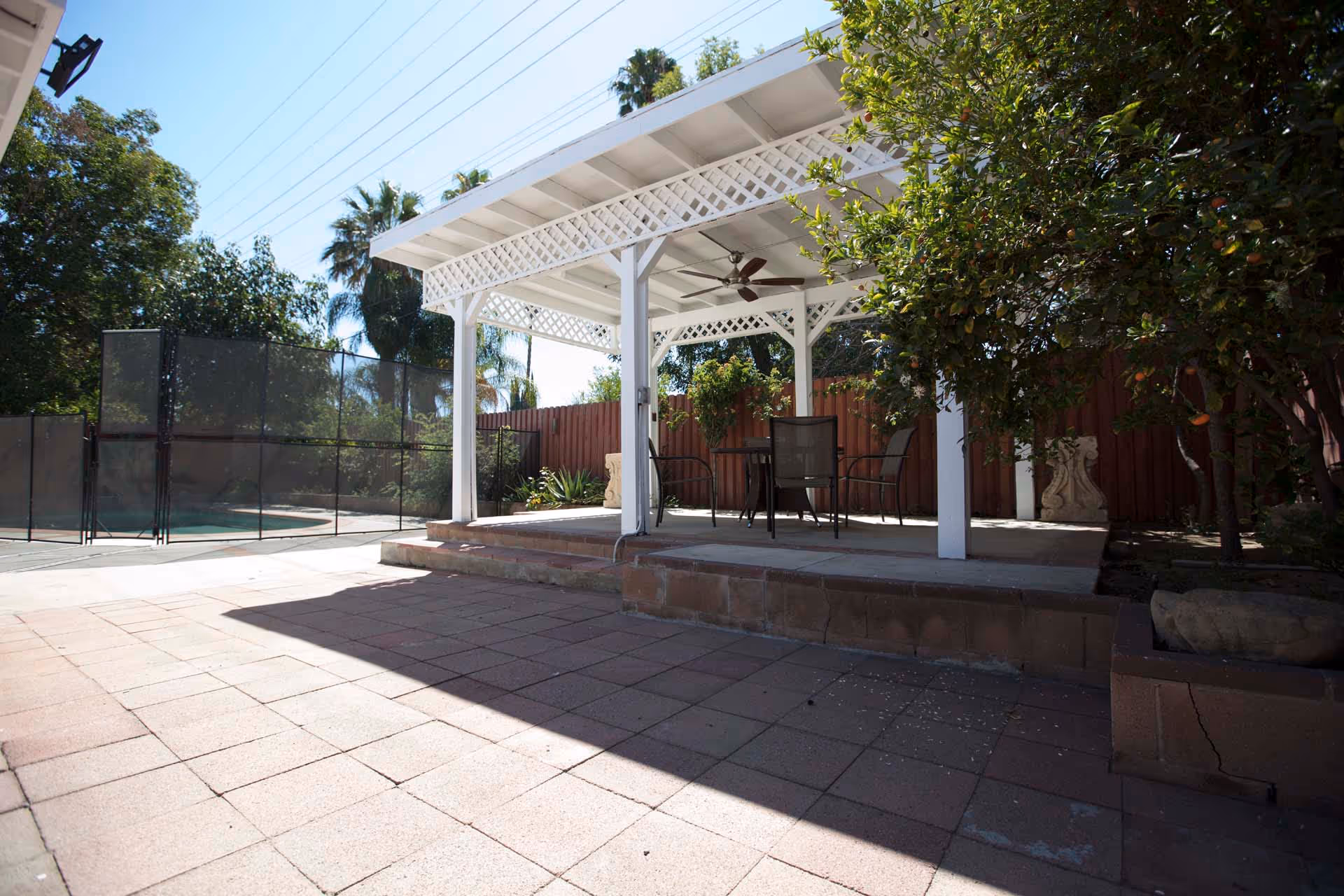 Outdoor patio area with a white pergola structure featuring lattice trim and a ceiling fan. Underneath the pergola are a table and chairs. The patio is paved with tiles and surrounded by trees and a wooden fence. A fenced pool area is visible in the background.
