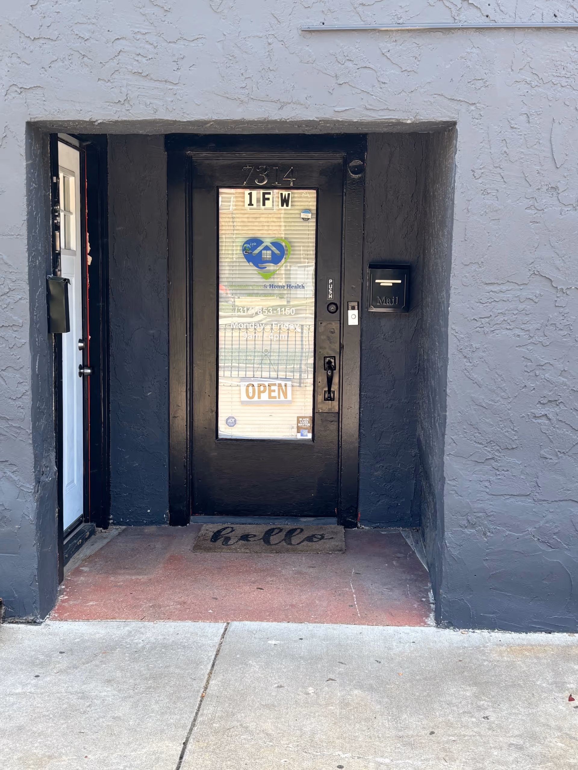 Black door entrance with glass panel, 'OPEN' sign, and welcome mat in gray stucco exterior.