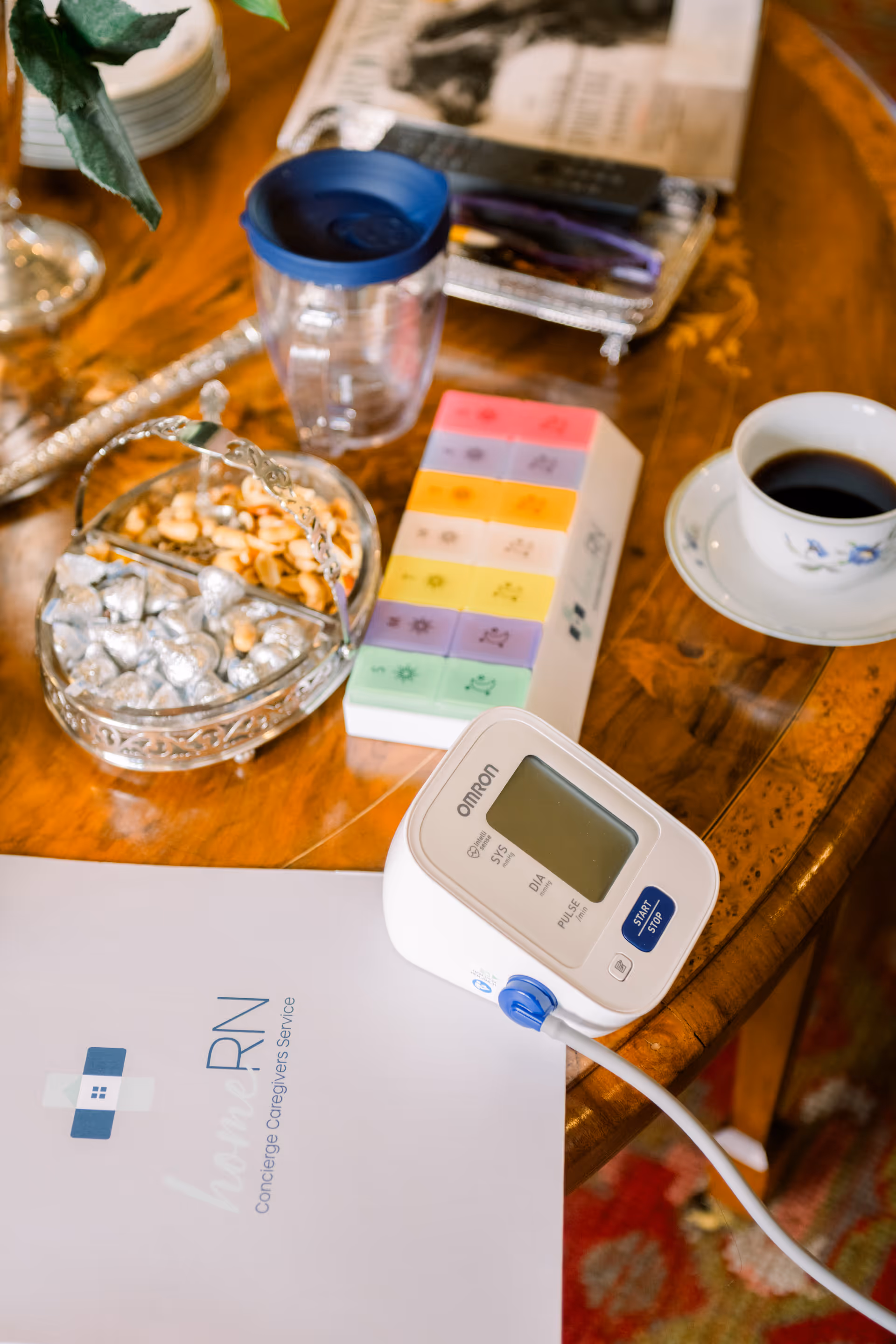 Interior table scene with an Omron blood pressure monitor, colorful pill organizer, coffee cup, and papers.