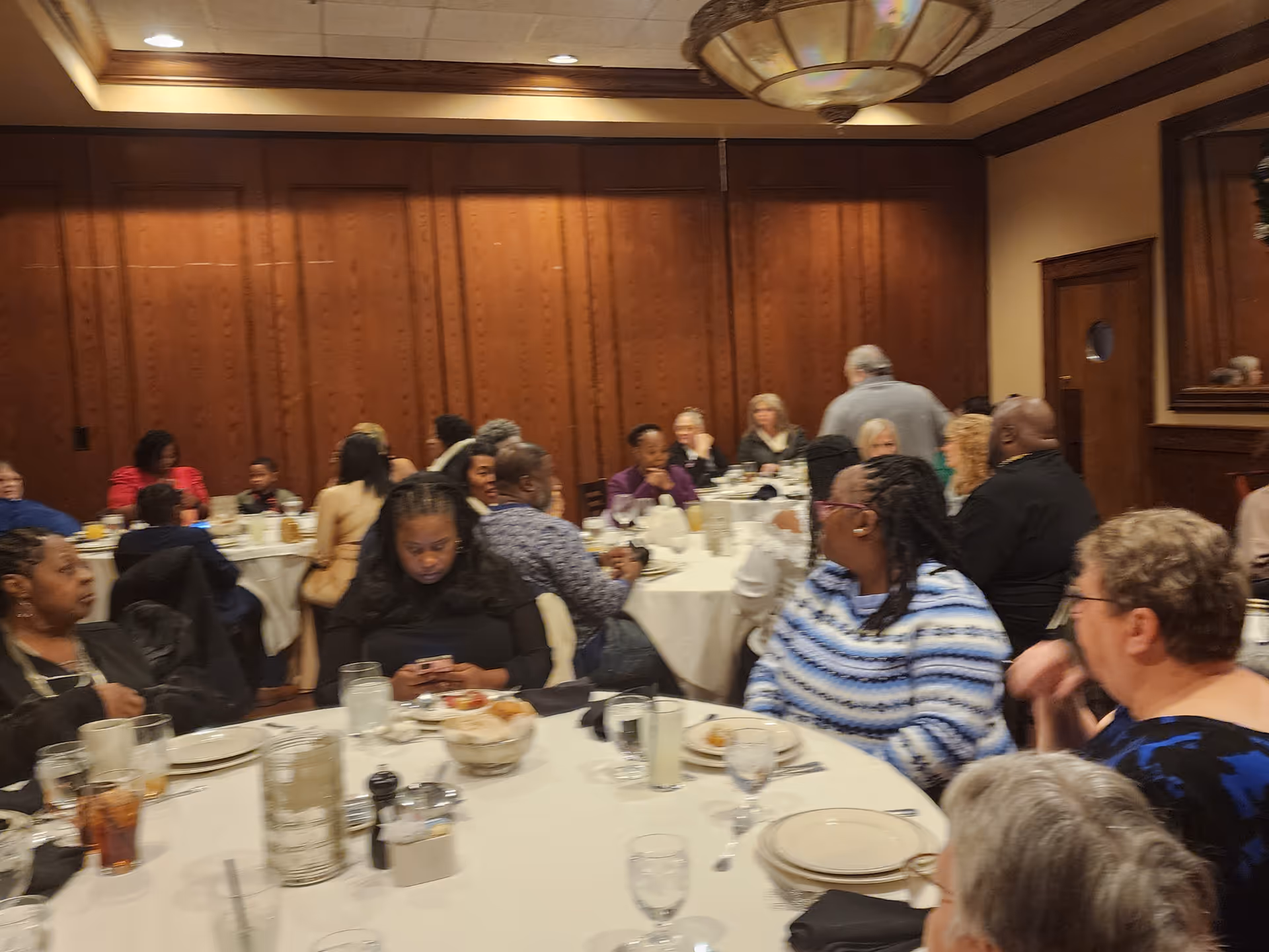 Residents seated at round tables in a wood paneled facility dining room.