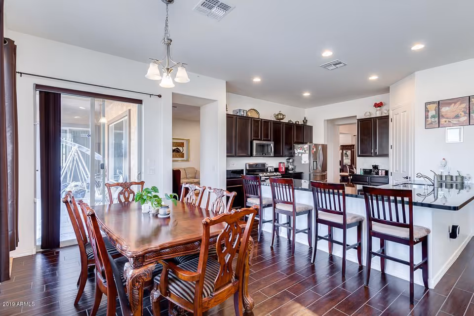 Bright and spacious kitchen and dining area with dark wooden cabinets, a granite countertop island with four high chairs, and a wooden dining table with six chairs. Sliding glass doors lead to an outdoor space, and the room features recessed lighting and a hanging light fixture above the dining table.
