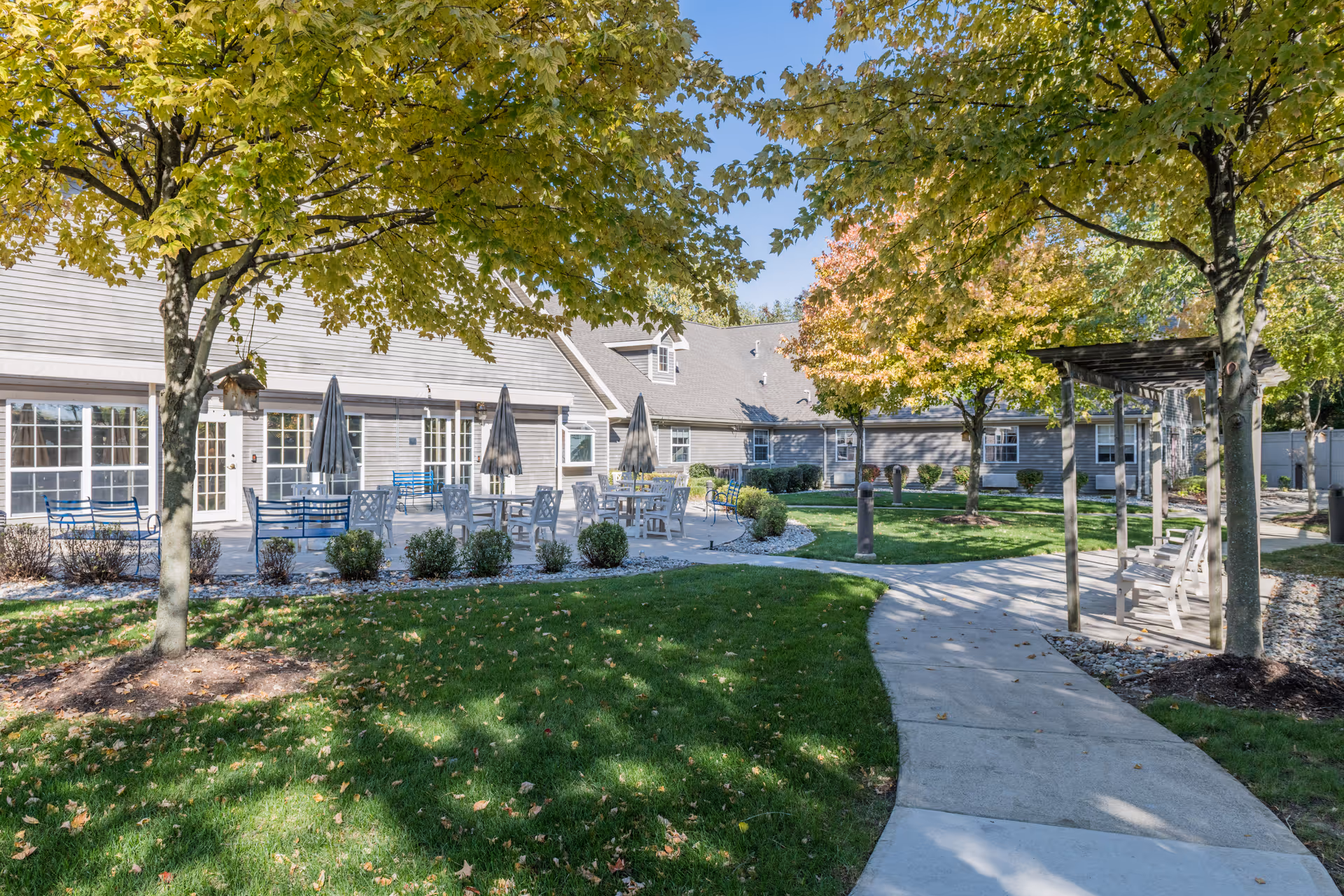 Outdoor courtyard of a memory care facility with patio tables and umbrellas, trees, a walkway, and a pergola.