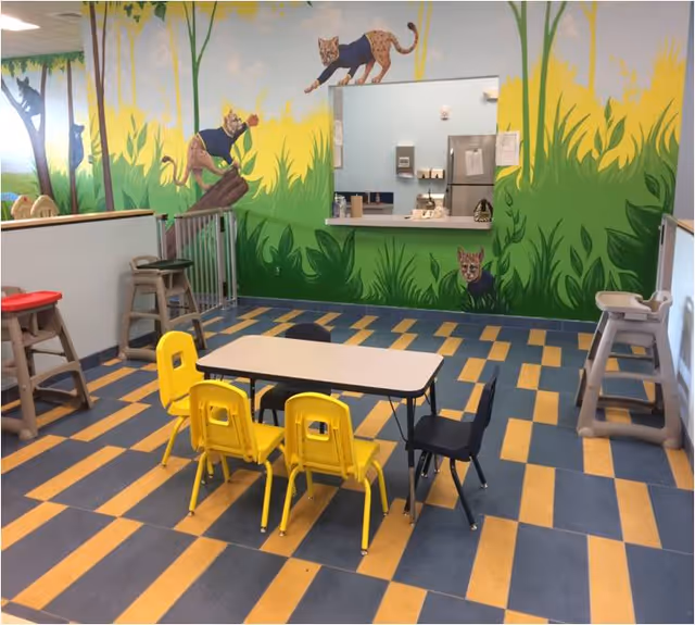 Bright indoor play area with a mural, small table, and kid-sized chairs.
