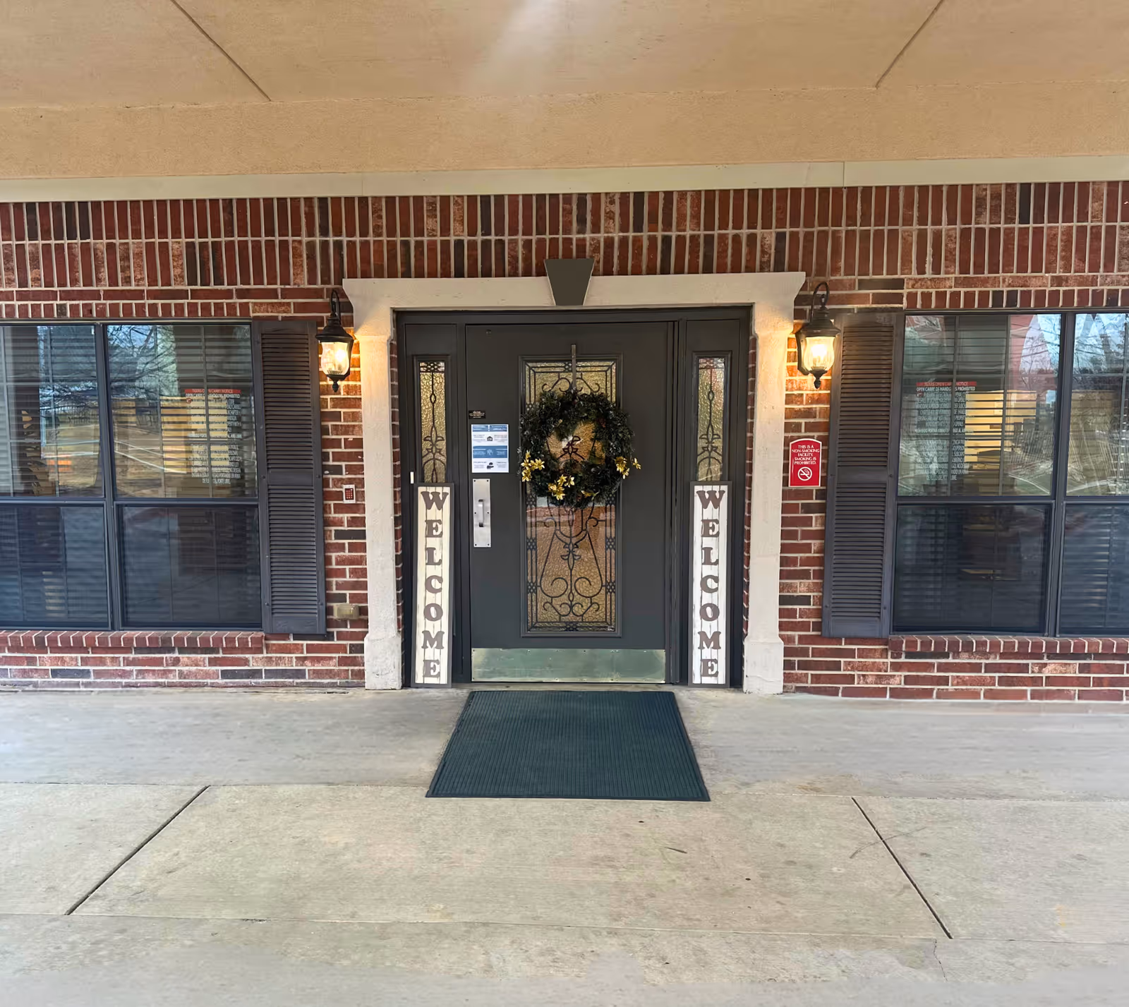Entrance door of a building with a decorative wreath hanging on it, flanked by two vertical signs that say 'WELCOME'. The door is framed by brick walls and two windows with dark shutters on either side. Two wall-mounted lantern-style lights are illuminated above the signs.