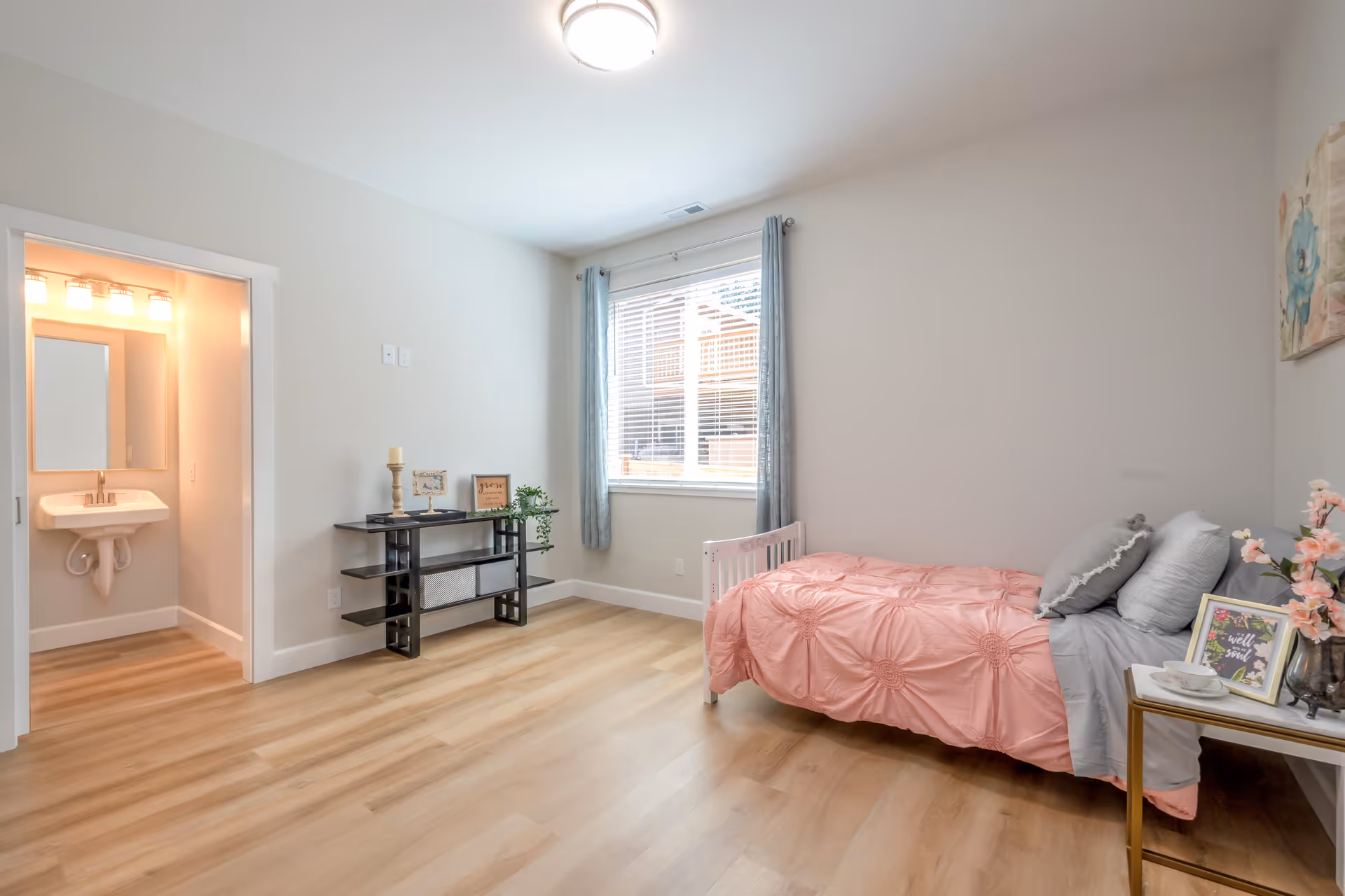 Bright bedroom with pink bedspread, grey pillows, window, and a bathroom doorway.